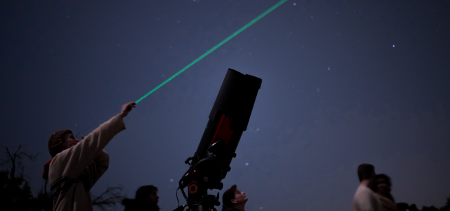 A person points to the night sky using a laser while standing next to a telescope.
