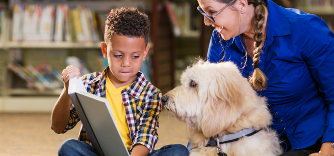 A young boy reads to a white dog while a smiling woman watches