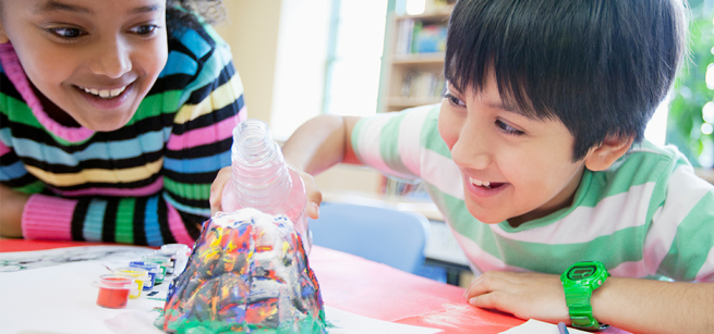 Two smiling children bent over a volcano science experiment pouring in the solution and waiting for it to bubble over.