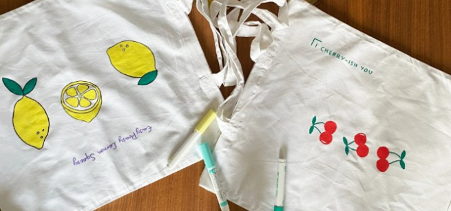 White canvas tote bags decorated with colorful fruit drawings and fabric markers on a table.