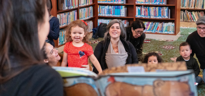 Babies and toddlers with grownups look to a person holding a book