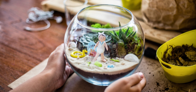Two hands holding a glass terrarium with a beach scene inside