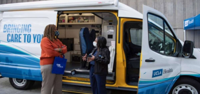 A white van with UCLA logo is parked with the passenger side door and sliding door open. Two women with medical masks stand outside the van, one is wearing medical scrubs. Inside the van shows medical equipment.