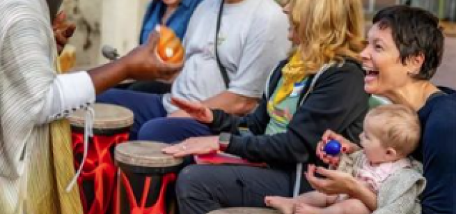 A musician holds a shaker egg instrument while people play drums and a caregiver holds a baby and shaker egg.