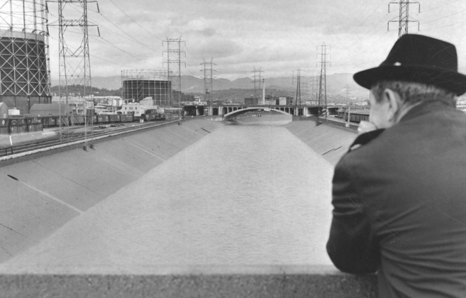 An unknown man gazes at the river under stormy skies, in a view replete with hulking oil tanks (known as “gasometers”) and other industrialized features. (1965, Herald Examiner Collection)
