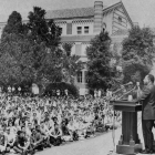 Martin Luther King Jr. standing in front of a podium on a stage, speaking to a large group of students who are sitting on the ground. 