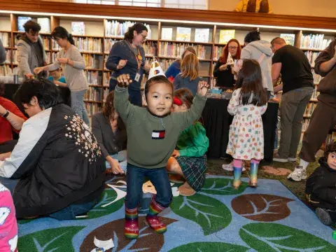 A toddler in a party hat dances with their arms up in a crowd of children and adults