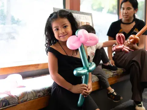 A young girl holds a balloon animal in front of a sunny window while a woman sits behind her.