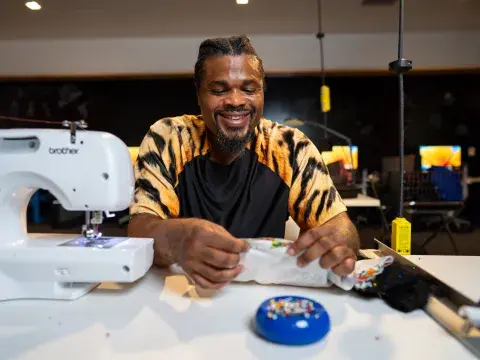 A man in tie-died shirt smiles while he works on a textile project at a white table with a sewing machine. 