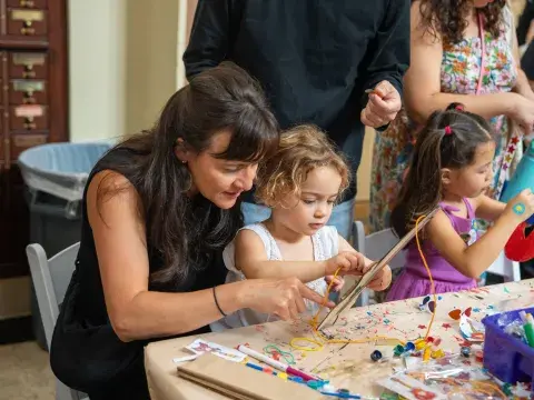 A woman and a young child bend over a table and work on a craft project in a crowded library setting.