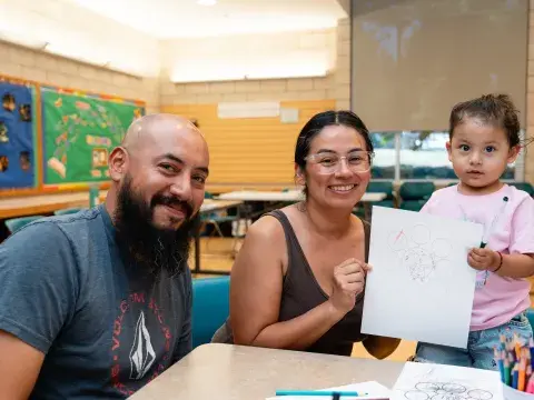 Two smiling adults sit at a table in a library next to a young child who is holding up a piece of artwork.