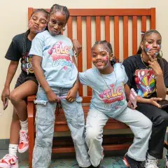 four children wearing facepaint sit on a wooden bench, posing and grinning for the camera.