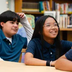 two children sit at a table in a library, smiling and watching something.