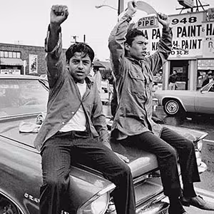 Two people sitting on the hood of the car during a Chicano rally.