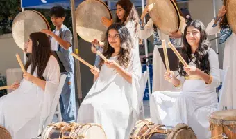 Three teenage girls in white gowns hold large drumsticks above large, ceremonial drums.