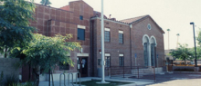 a two story mostly red brick library building in a paved parking lot area with two trees on the left side and a flag pole