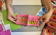 hands displaying pop up book art project, scissors and paper on table