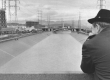 A black and white photograph with a man in overcoat and wide-brimmed hat facing away from the camera, looking down at the Los Angeles River with industrial tanks in the background.