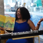A musician belts out a song while reading a storybook to children. She sits at a musical keyboard and is surrounded by books.