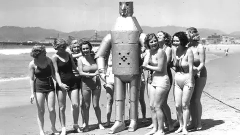 Robot on the beach surrounded by ladies in bathing suits