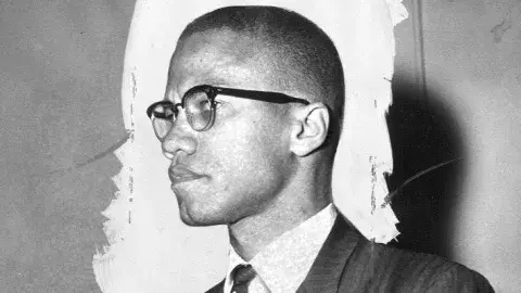 Black and white photo of Malcolm X from the side, standing at a meeting.