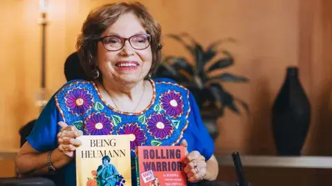 Activist Judy Heumann holding her two books, Being Heumann: An Unrepentant Memoir of a Disability Rights Activist and Rolling Warrior