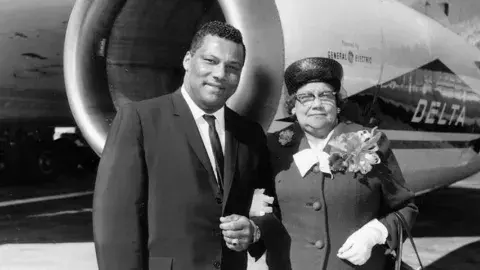 Photographer Rolland J. Curtis and his mother, Mathilda Curtis. They are standing near a Delta Airlines plane, and she is wearing a corsage.
