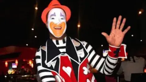 An adult clown waving his left hand.
