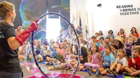 Adult holding a hula hoop, making bubbles, in front of a crowd of children