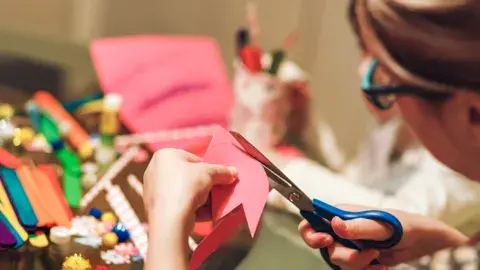 Adult woman cutting pink craft paper with scissors.