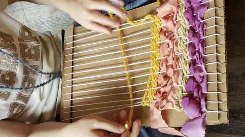 hands weaving yarns and ribbons on a cardboard loom
