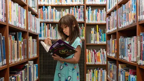 an elementary school age girl stands in a library aisle, surrounded by shelves of books, and reading an open book in her hands