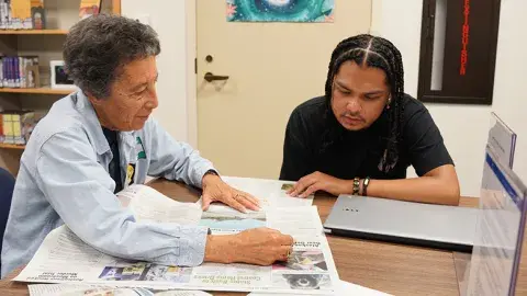 A person helping another person read a newspaper.