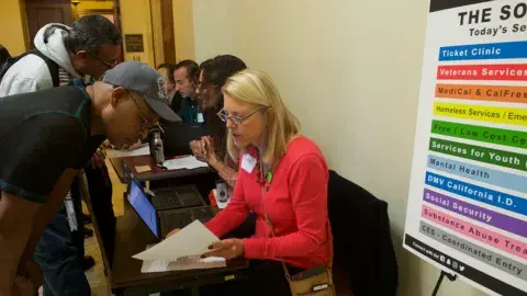 Image of a seated woman in a red sweater at a table helping a standing man with paperwork.