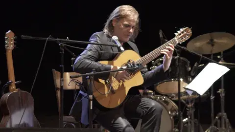 Fabio Zini is shown mid-performance playing a nylon string guitar wearing a shiny black suit. 