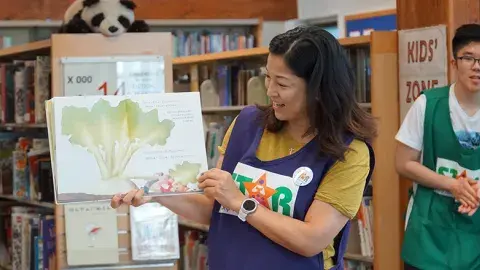 Smiling person wears a purple apron with the word STAR on it. The person is standing in front of library shelves and holding up a picture book. They appear to be reading aloud from it as they turn the pages. A similarly attired person stands nearby.