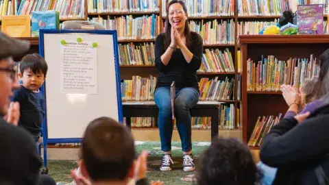 A woman sitsin front of a group of toddlers and adults clapping her hands. A white board is next to her facing the crowd. 