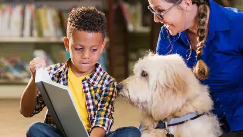 A young boy, cozily sitting in the library, reading to a BARK Therapy Dog and the dog's happy owner.
