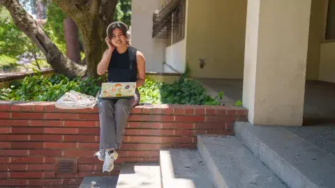 A teenage girl sits on a brick wall next to a staircase. She is using a laptop covered with stickers and wearing headphones and smiling.