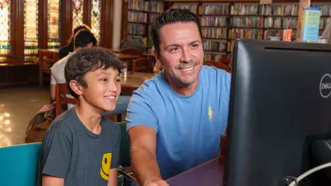 A man and a boy use a computer in a library.