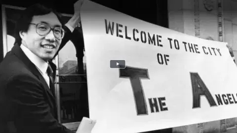 historic black and white photo of man holding sign "welcome to the city of the angels"