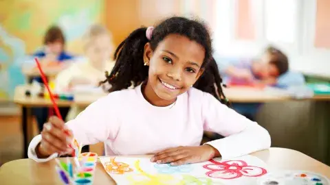 A smiling, young girl uses a paintbrush and colored paint pots to create a watercolor illustration of flowers.