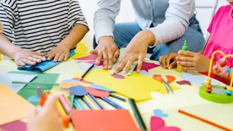 Children and an adult gather around a table and create art using a variety of materials. 