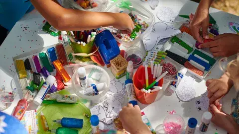 An assortment of craft supplies, paints, markers and hands with scissors cutting paper.