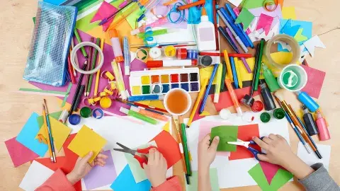 An assortment of craft supplies, paints, markers and hands with scissors cutting paper.