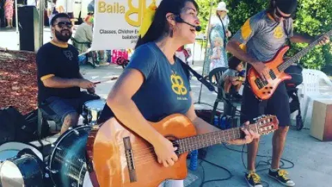 Performer Isabel Brazon playing guitar with band outdoors.
