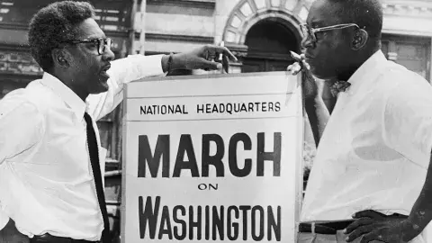 Bayard Rustin at a sign about the March on Washington talking with another man.