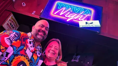 The hosts of It Wonders Me Trivia next to a picture of their logo of a covered wagon silhouette 