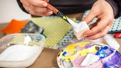 A table with paint supplies and a pair of hands painting a small block of wood.