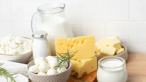 Assorted dairy products on a wooden surface, including milk in a jug, various cheeses, yogurt, and cream with sprigs of rosemary, against a tiled background.